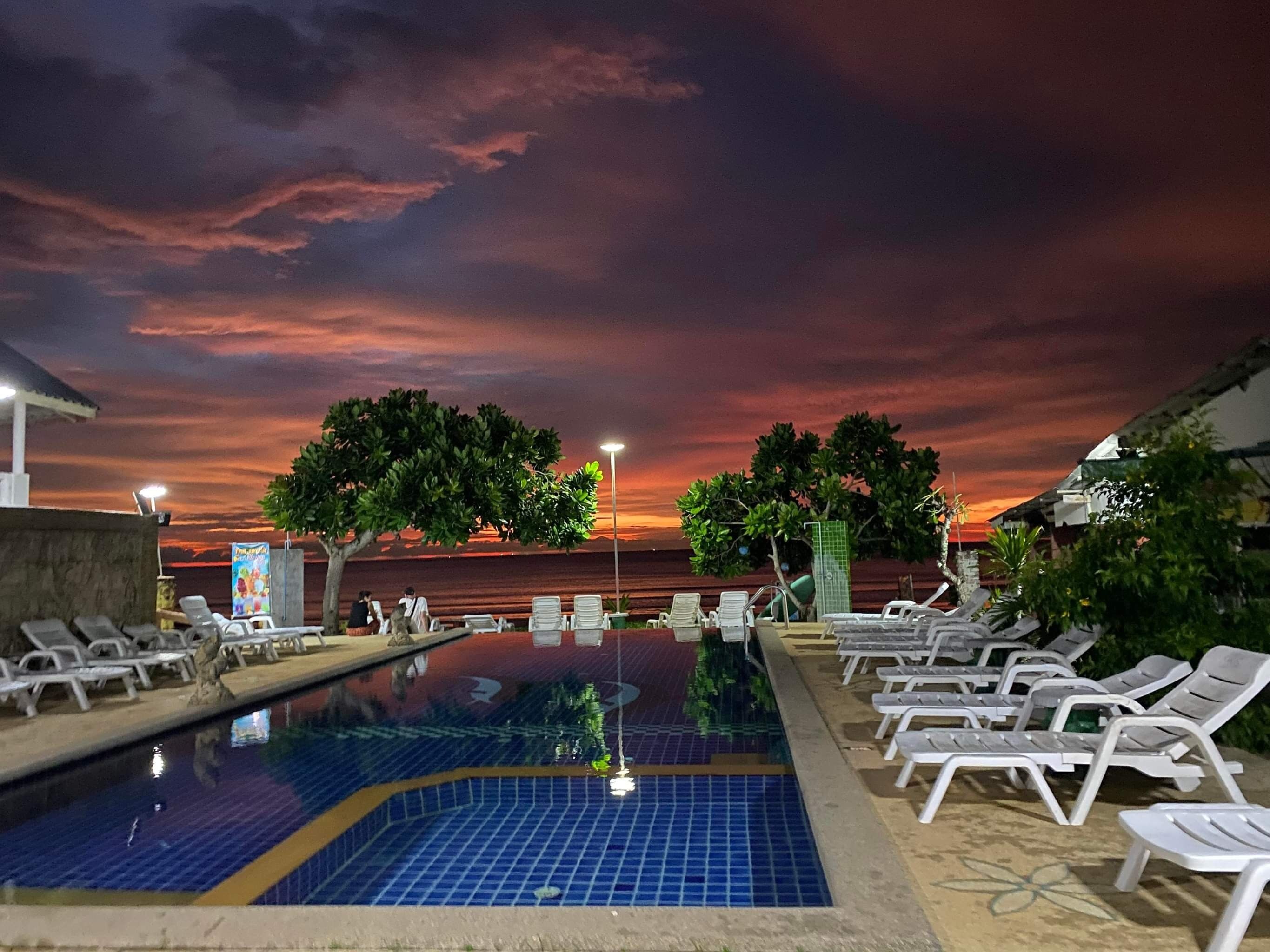 Piscina con sillones de descanso y árboles, frente a un cielo al atardecer.