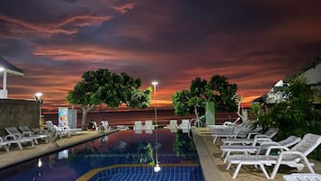 Piscina con sillones de descanso y árboles, frente a un cielo al atardecer.