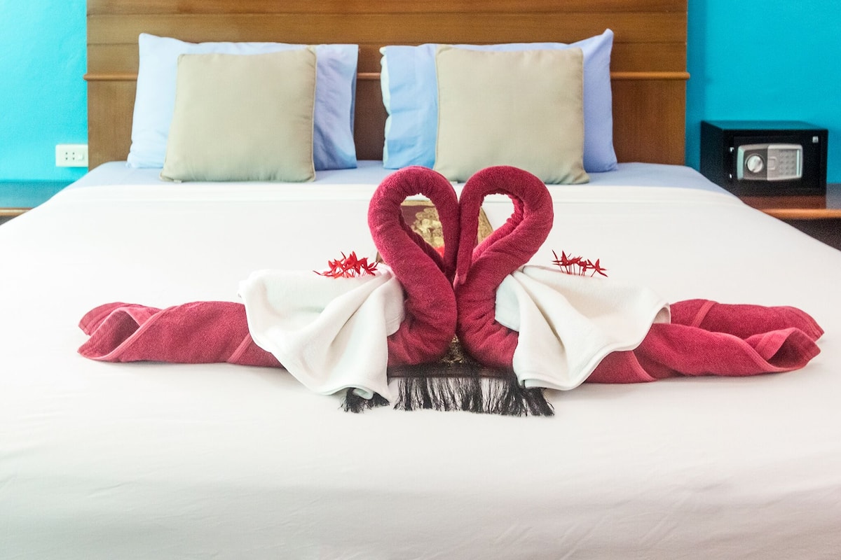 A neatly made bed with two red towels arranged in a heart shape, white pillows, and a small black radio on the nightstand.