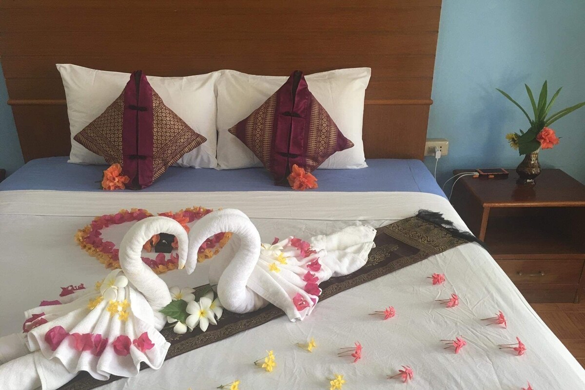 A hotel room with a neatly made bed, two pillows, a nightstand with a vase of flowers, and a decorative swan towel arrangement.