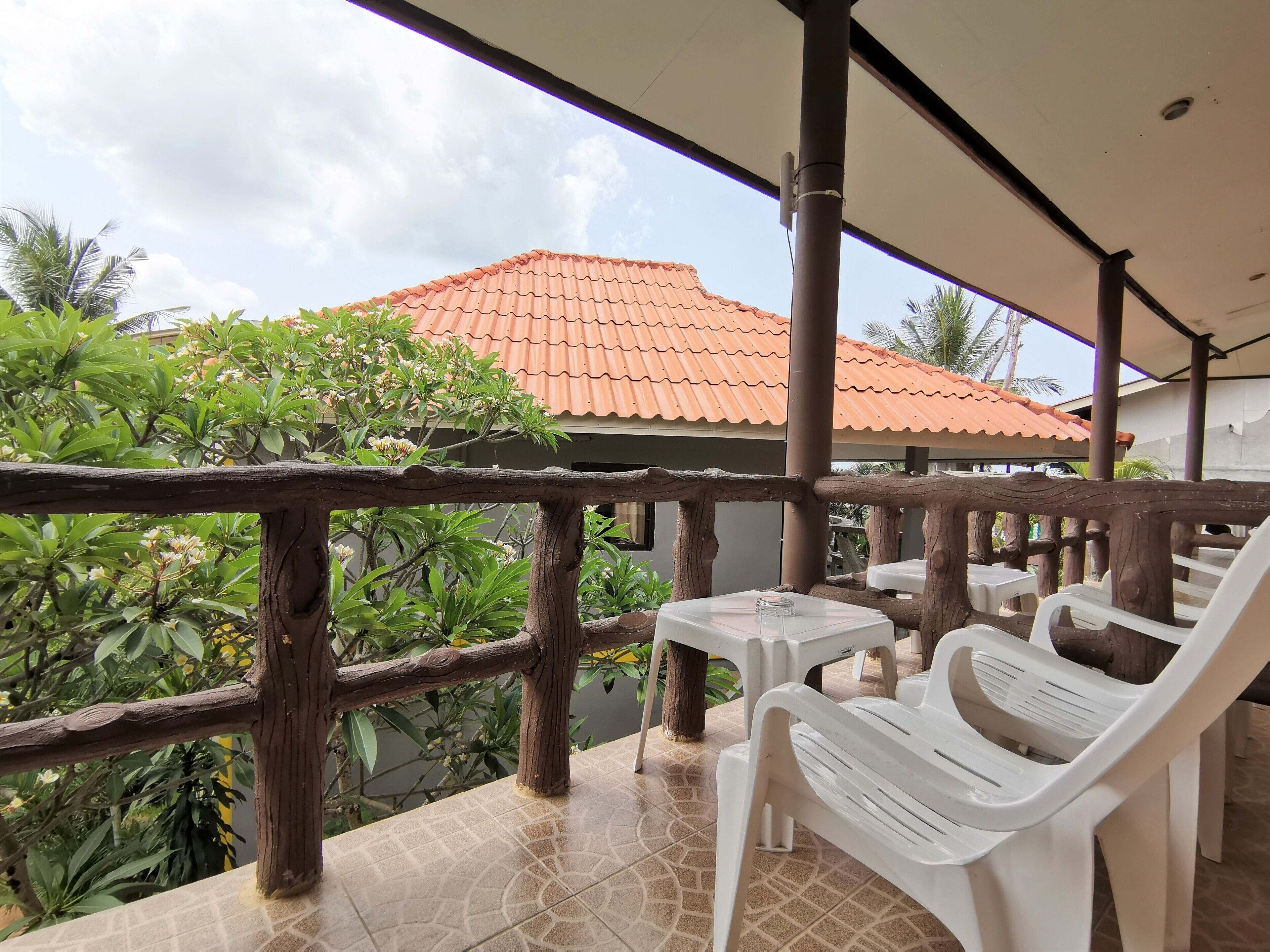 A covered outdoor seating area with white plastic chairs and tables, a wooden railing, and a view of a house with a red-tiled roof.