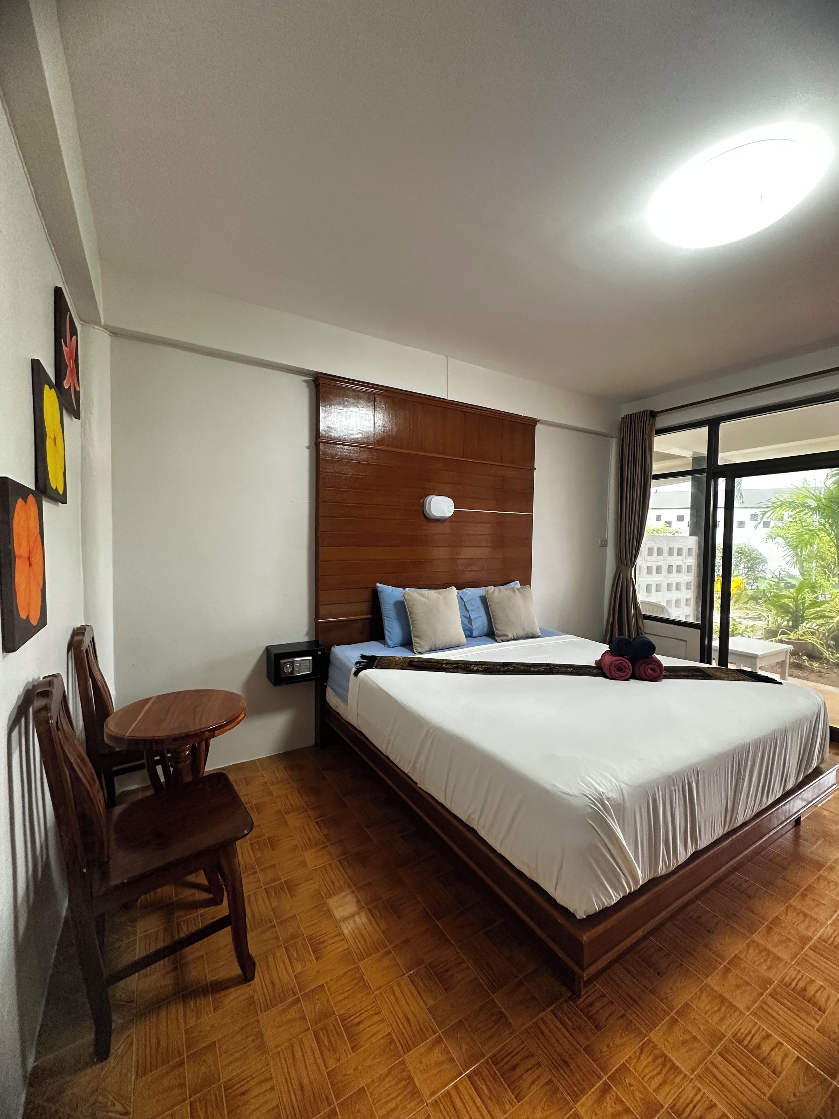 Habitación de hotel con cabecera de madera, cama con ropa de cama blanca, una mesa de comedor de madera con sillas y un ventanal con vistas a edificios y vegetación.