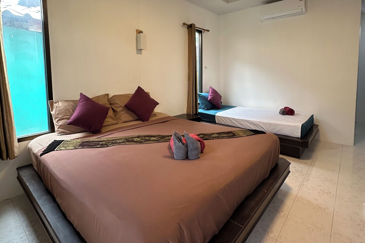 A hotel room with a large bed, a smaller bed, a window with curtains, an air conditioning unit, and a ceiling light.