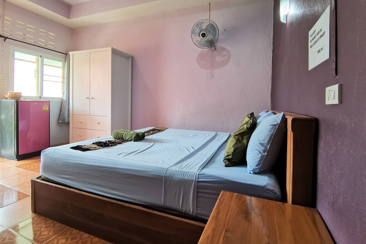 A bedroom with a bed, a fan, a wardrobe, and a small table.
