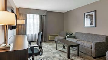 A hotel room with a sofa, a small table, a chair, a desk with lamps, a TV, and a framed picture on the wall.