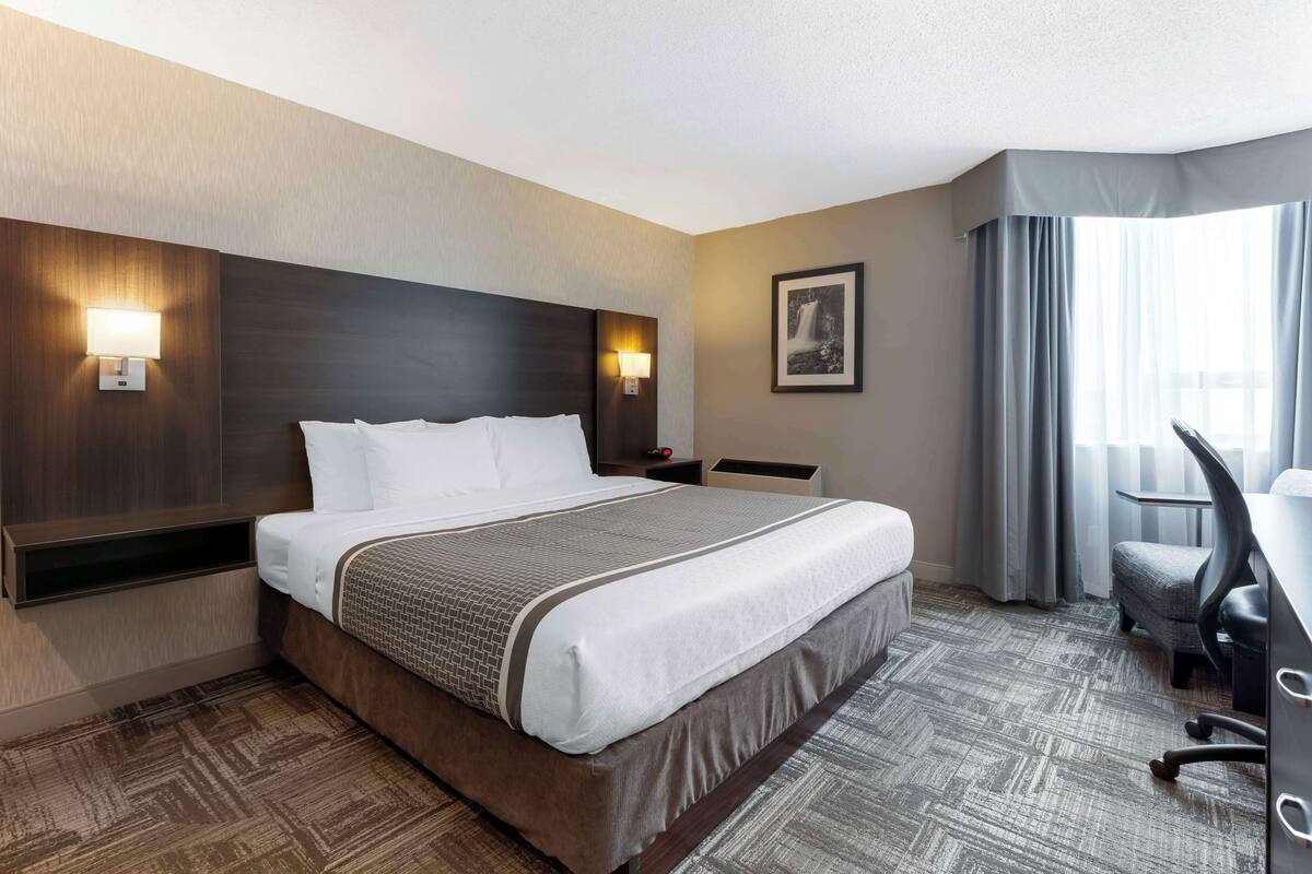 Habitación de hotel con una cama grande, dos mesitas de noche con lámparas, un escritorio con silla, una ventana con cortinas e imagen enmarcada en la pared.