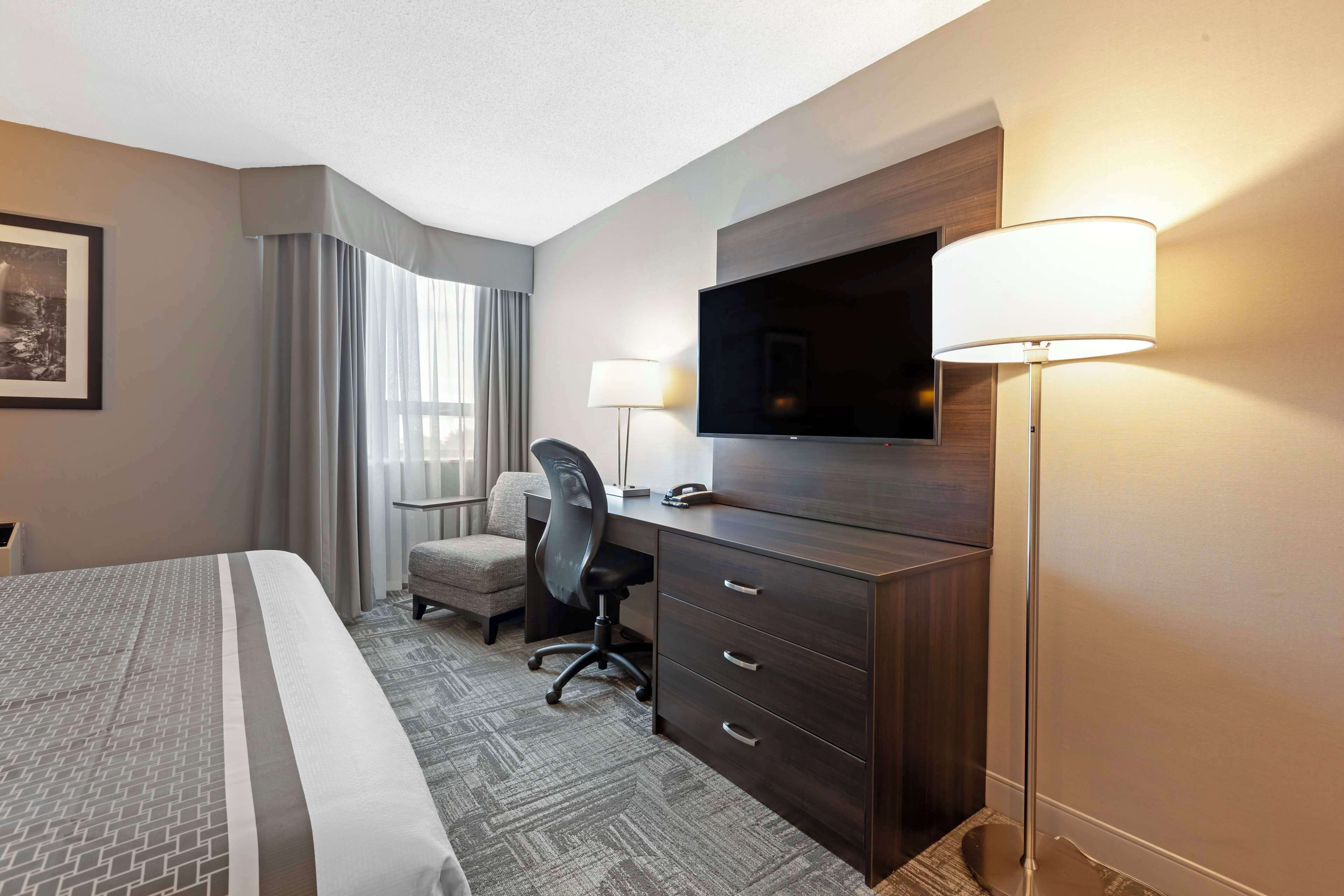 Quarto de hotel com cama, escrivaninha, cadeira, televisão e abajur.