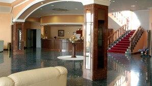Un'ampia hall con pavimento in marmo lucido, una reception, una scala rivestita di tappeto rosso e un divano beige.