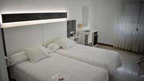 Una camera d'albergo con due letti, una scrivania, una sedia, una lampada e una finestra con tende.