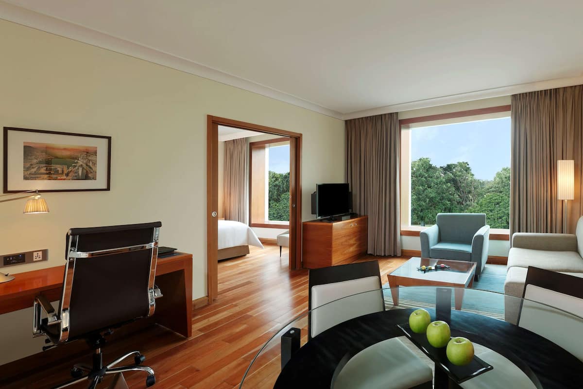 A hotel room with a wooden desk, a comfortable sofa, a glass coffee table with apples, and a TV on a wooden cabinet.