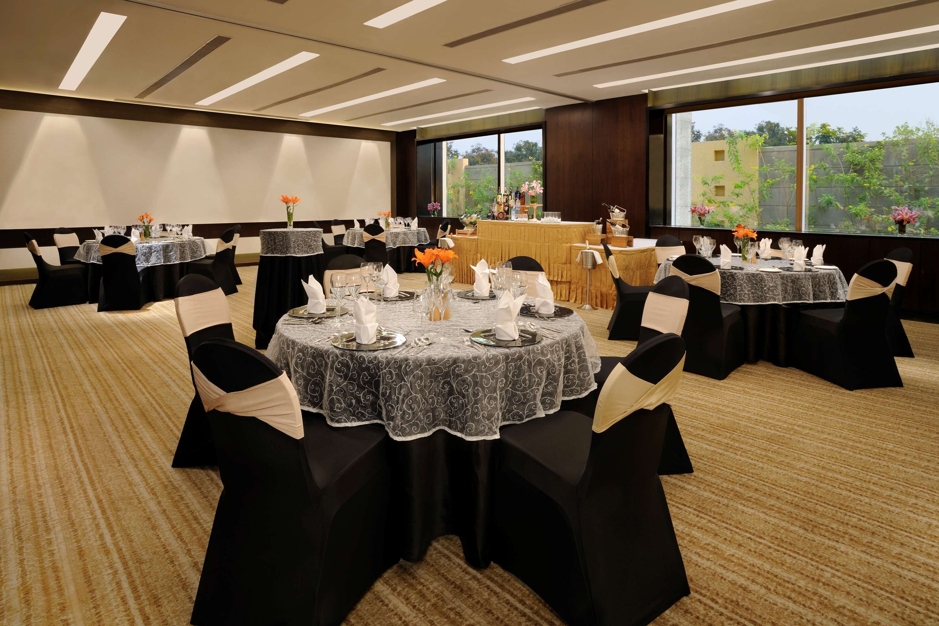 Une salle de conférence dotée de plusieurs tables rondes, chacune agrémentée de serviettes blanches, de verres et de fleurs oranges. La salle est recouverte d’un tapis et possède de grandes fenêtres offrant une vue sur la verdure environnante.