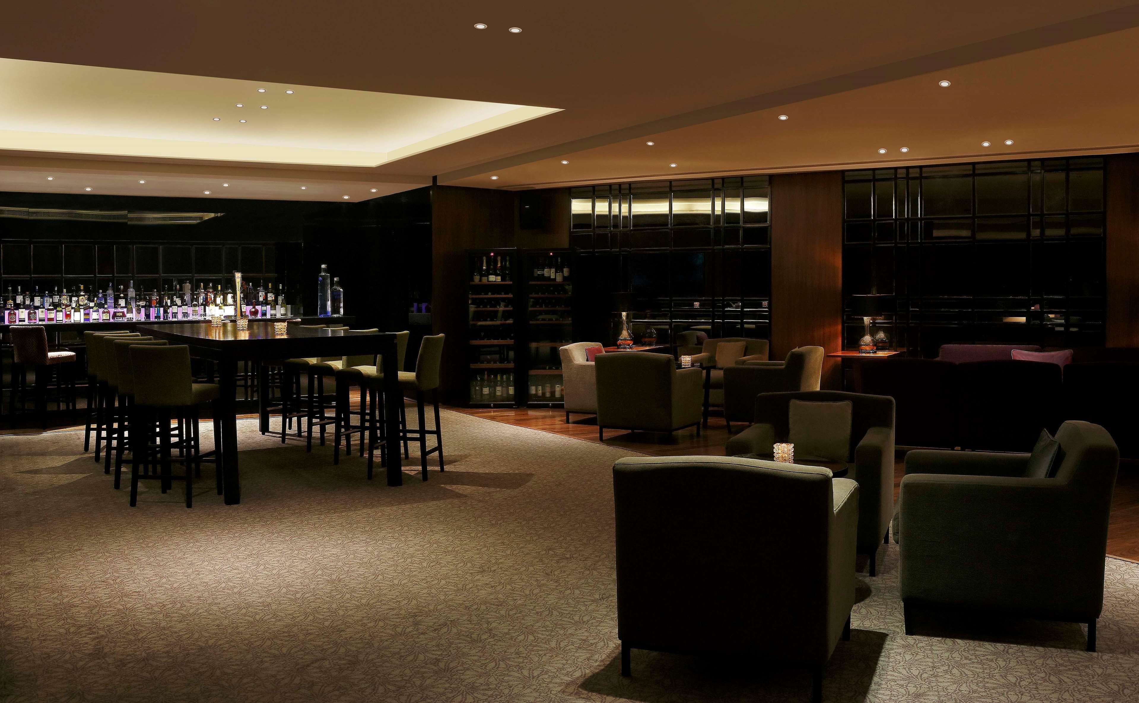 A dimly lit bar area with a long counter, multiple chairs, and shelves stocked with bottles.