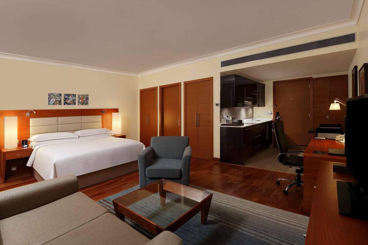 A hotel room with a bed, a desk, a chair, a sofa, a TV, and a kitchenette.
