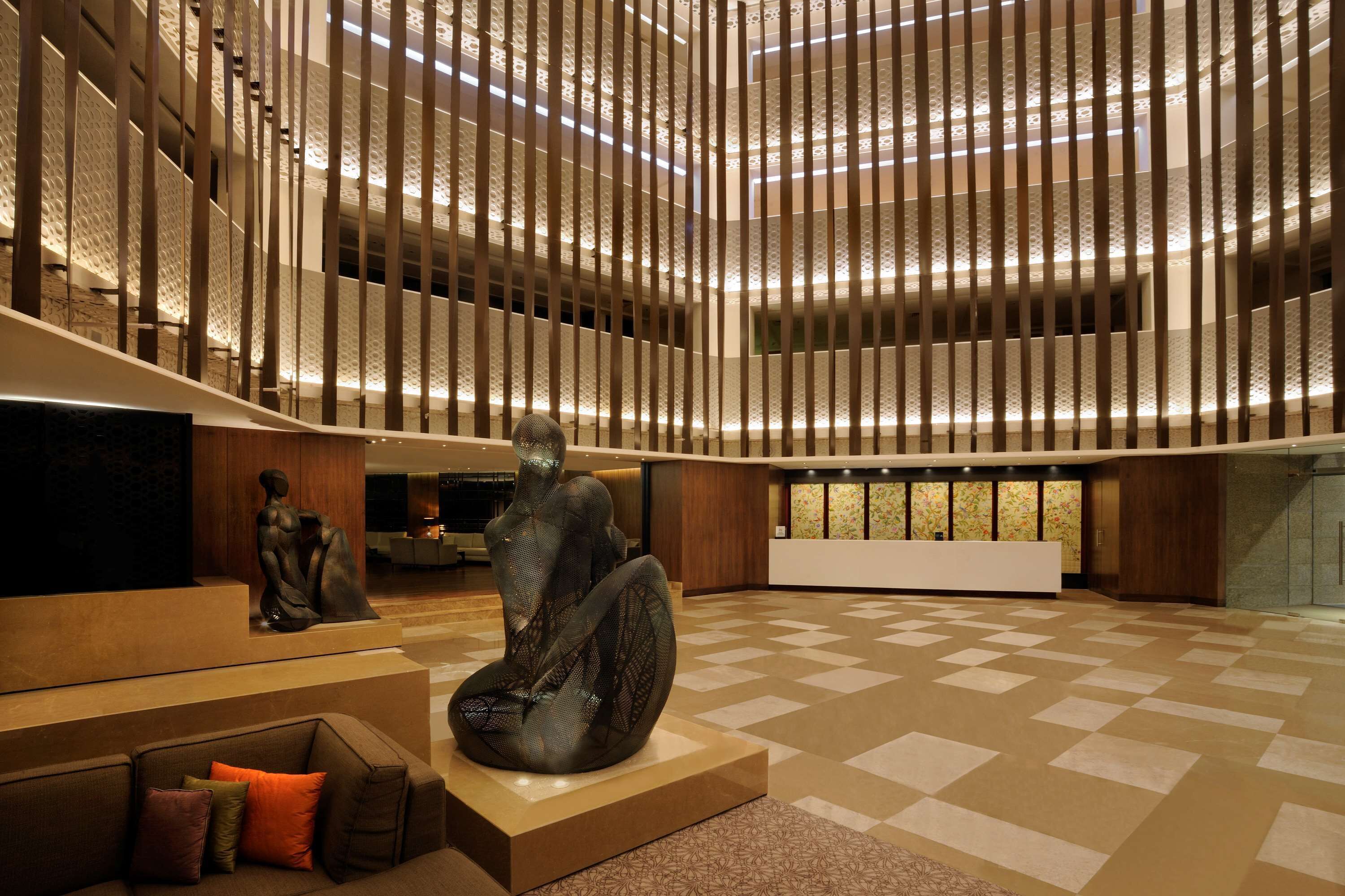 A spacious lobby with a modern design, featuring a central sculpture, a seating area with a sofa and cushions, and a checkered floor pattern.