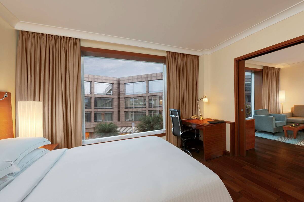 A hotel room with a large bed, a desk with a chair, a TV, and a view of a building through the window.