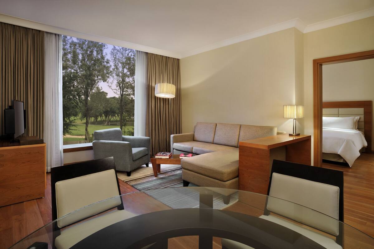 A hotel room with a sofa, armchairs, a dining table, a TV, a lamp, and a bed.