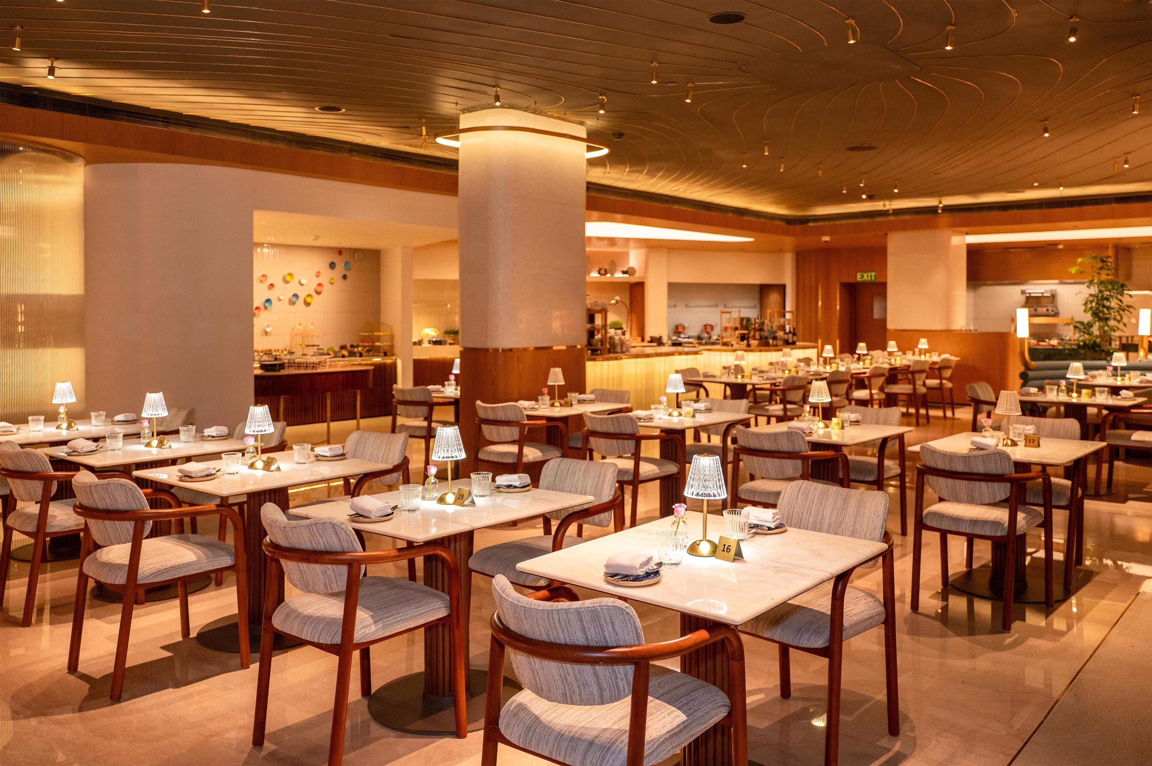 A spacious hotel dining area with multiple tables and chairs, a central pillar, and a bar area in the background.