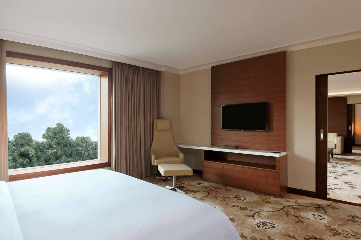 A hotel room with a large bed, a desk, a chair, a TV, and a view of trees through the window.