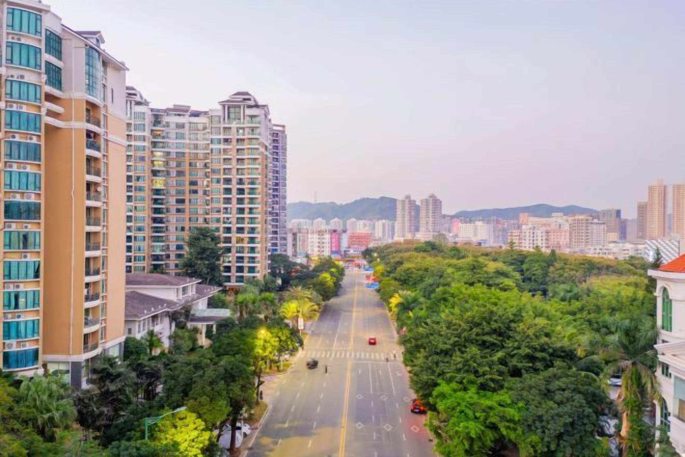 A multi-story residential building with a central road, surrounded by greenery and other buildings in the background.