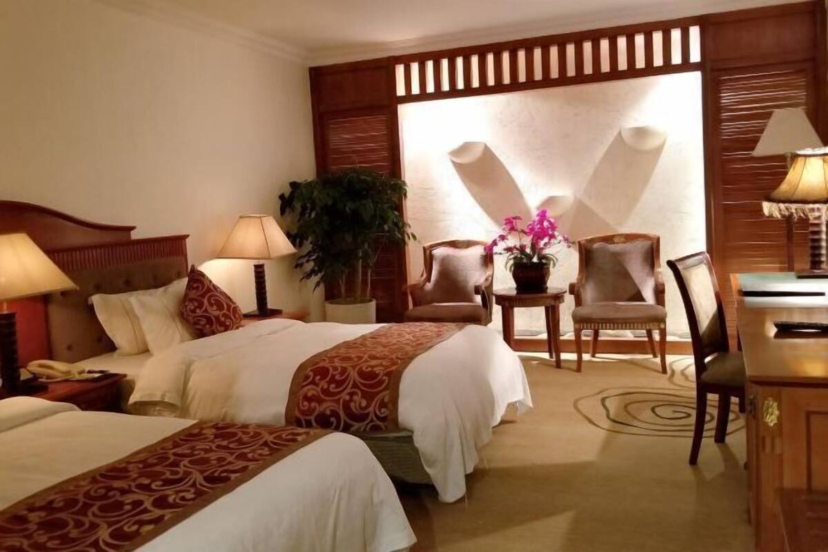 Habitación de hotel con dos camas, un escritorio, una silla, una lámpara, una planta y un ventanal grande.
