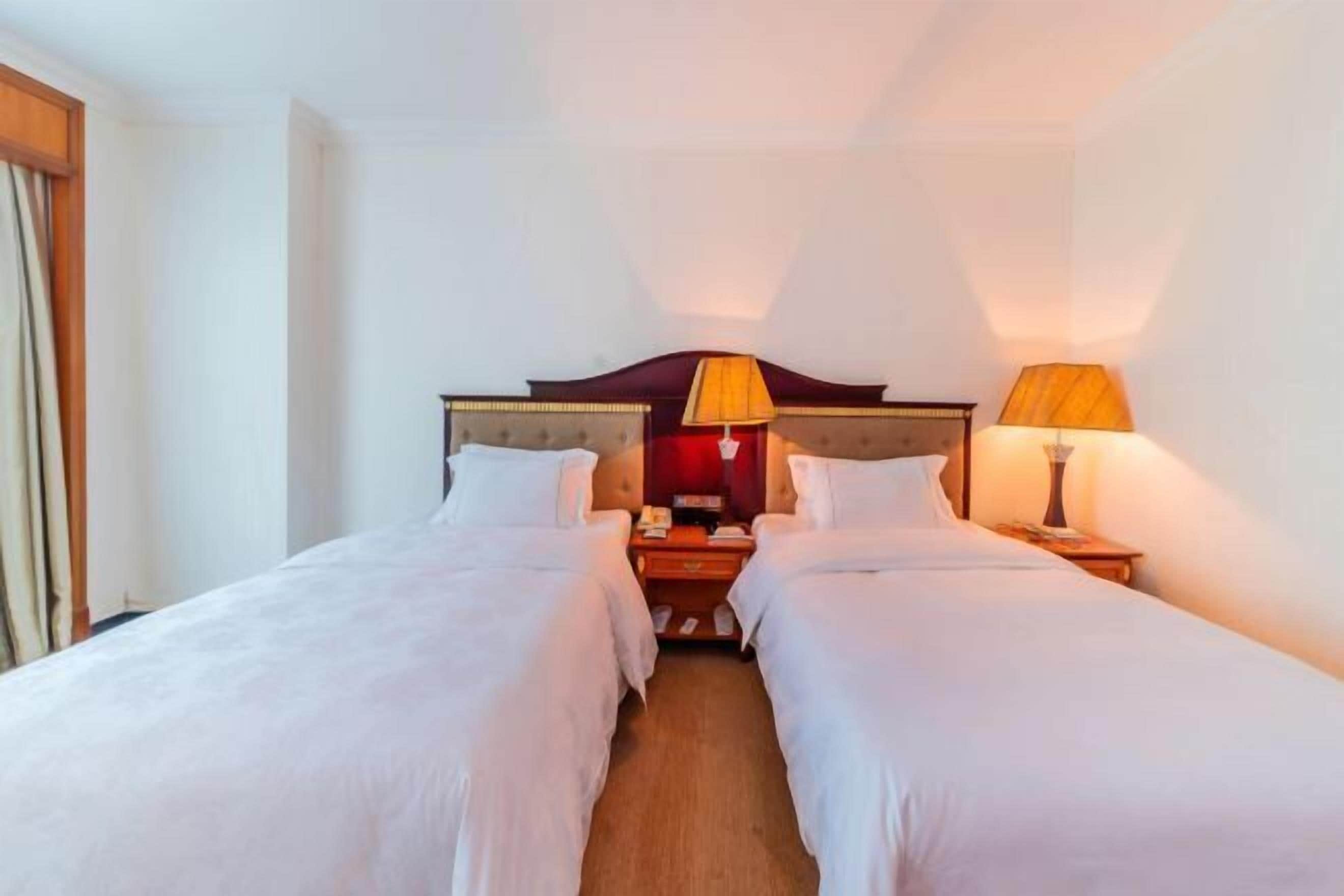 A hotel room with two single beds, a wooden headboard, bedside tables with lamps, and a nightstand.