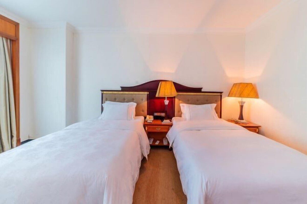 A hotel room with two single beds, a wooden headboard, bedside tables with lamps, and a nightstand.