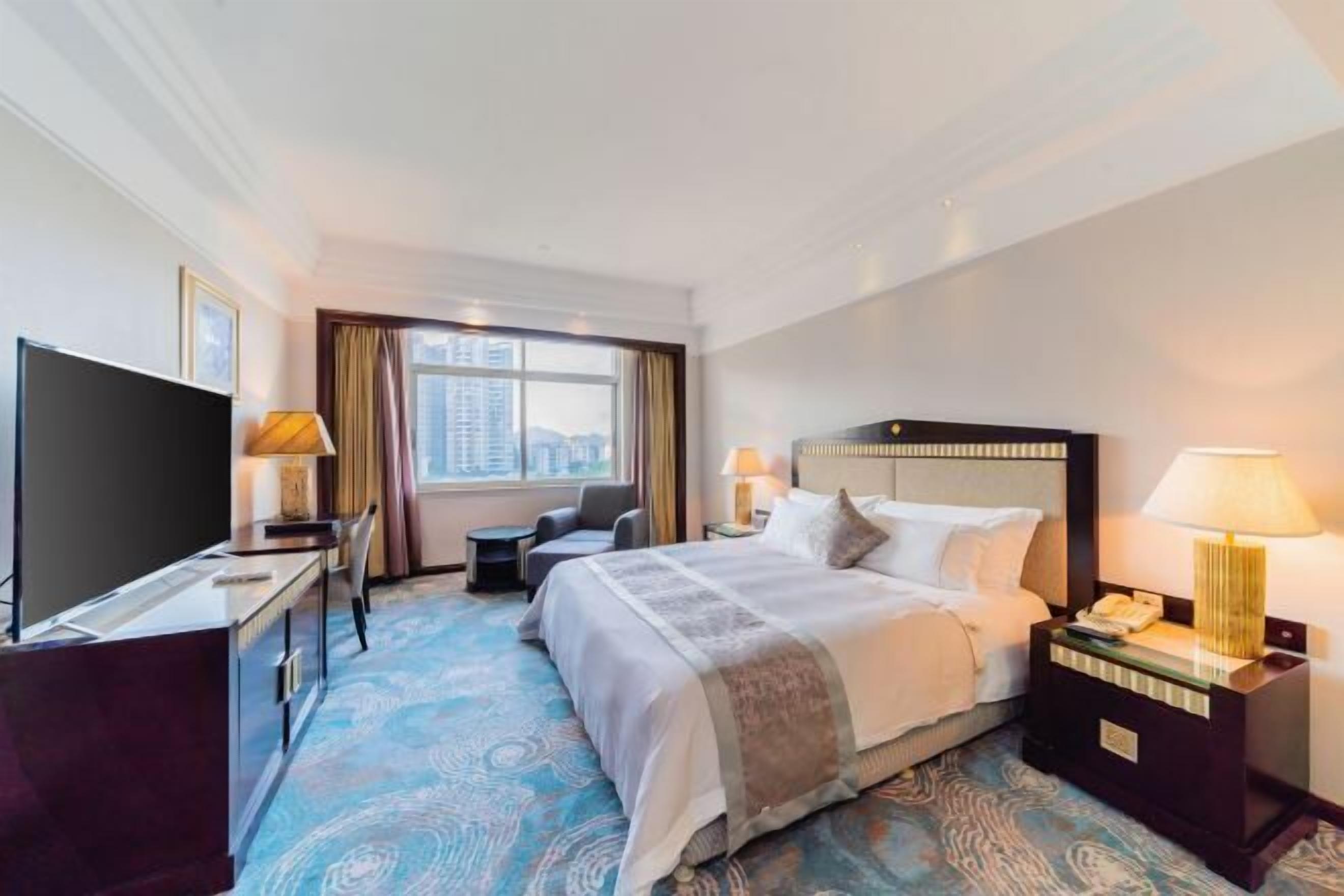 A hotel room with a bed, a desk with a TV, a chair, a nightstand with a lamp, and a view of the city.