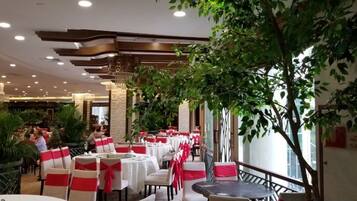 Un restaurante con mesas y sillas redondas, algunas con fajas rojas, y un árbol grande en el centro.