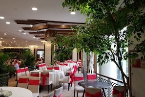 Un restaurant avec des tables et des chaises rondes, dont certaines sont ornées de rubans rouges, et un grand arbre au centre.