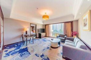 A hotel room with a large window, a sofa, a TV, a desk with a lamp, and a patterned rug.