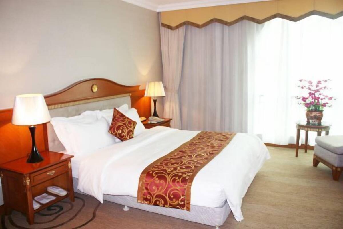 Habitación de hotel con cama, dos mesitas de noche con lámparas, una pequeña mesa con un jarrón de flores y una ventana con cortinas.