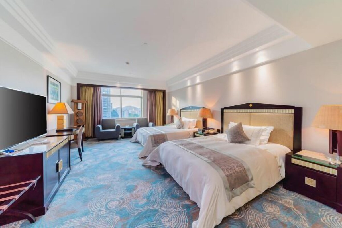 A hotel room with two beds, a desk, a TV, and a large window with a city view.