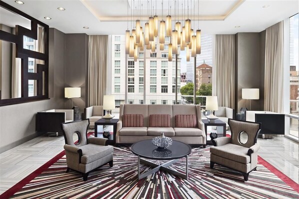 A modern hotel lobby with a central sofa, two armchairs, a coffee table, and a chandelier. The room features a large window with a city view.