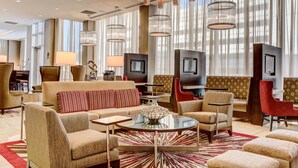 Um amplo hall de hotel com design moderno, apresentando uma área de estar central com sofás e poltronas, uma mesa de centro e uma televisão.