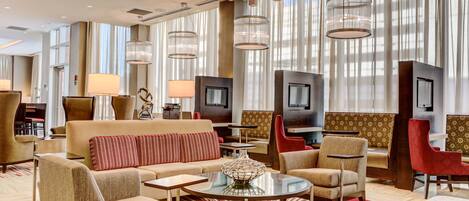 A spacious hotel lobby with a modern design, featuring a central seating area with sofas and armchairs, a coffee table, and a television.
