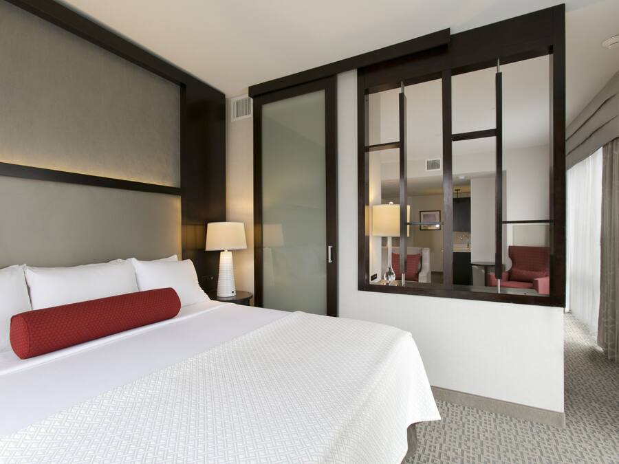 A hotel room with a large bed, a red pillow, a nightstand with a lamp, a sliding glass door, and a view of another room.