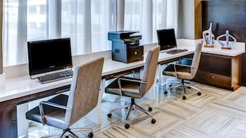 A modern office space with three desks, each with a laptop and a printer. The room has a herringbone floor pattern and large windows with sheer curtains.