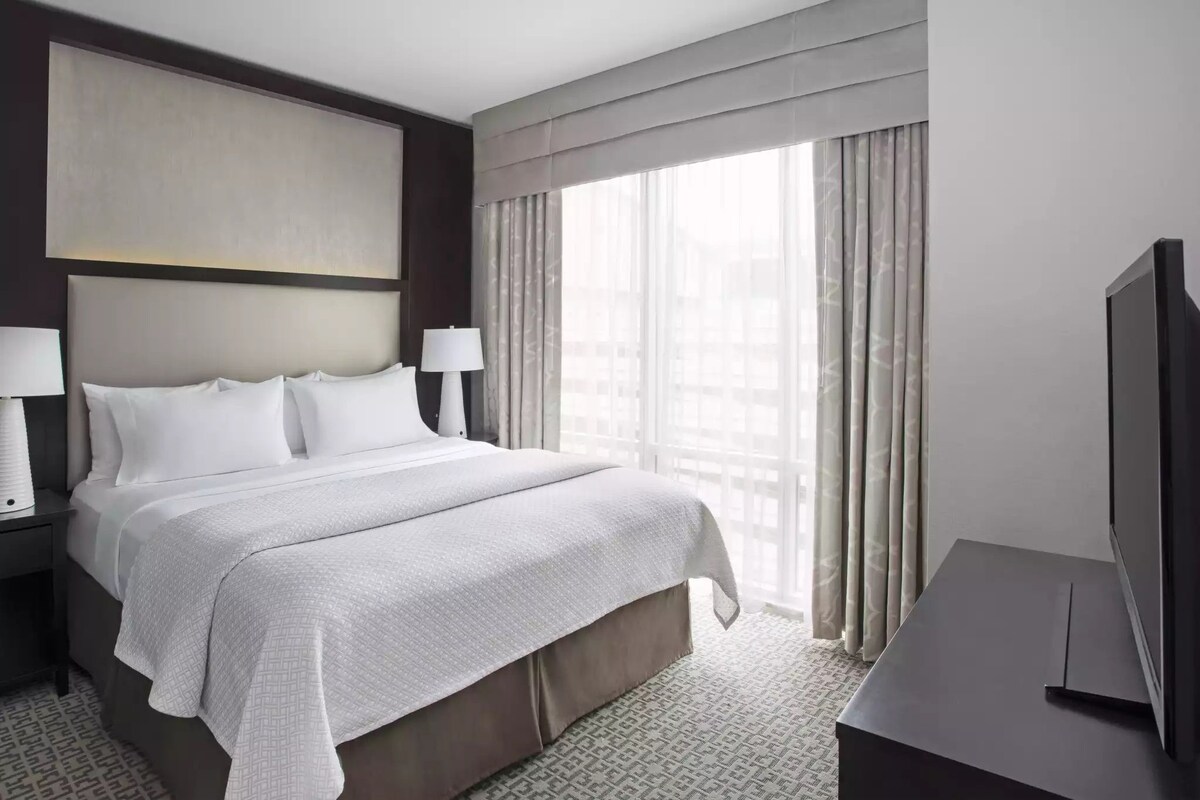 A hotel room with a large bed, two bedside tables with lamps, a flat-screen TV, and a large window with curtains.