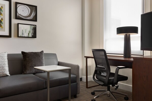 Una habitación de hotel moderna con un sofá de cuero gris, una mesita blanca, una silla de oficina negra, un escritorio y una lámpara de pie grande.