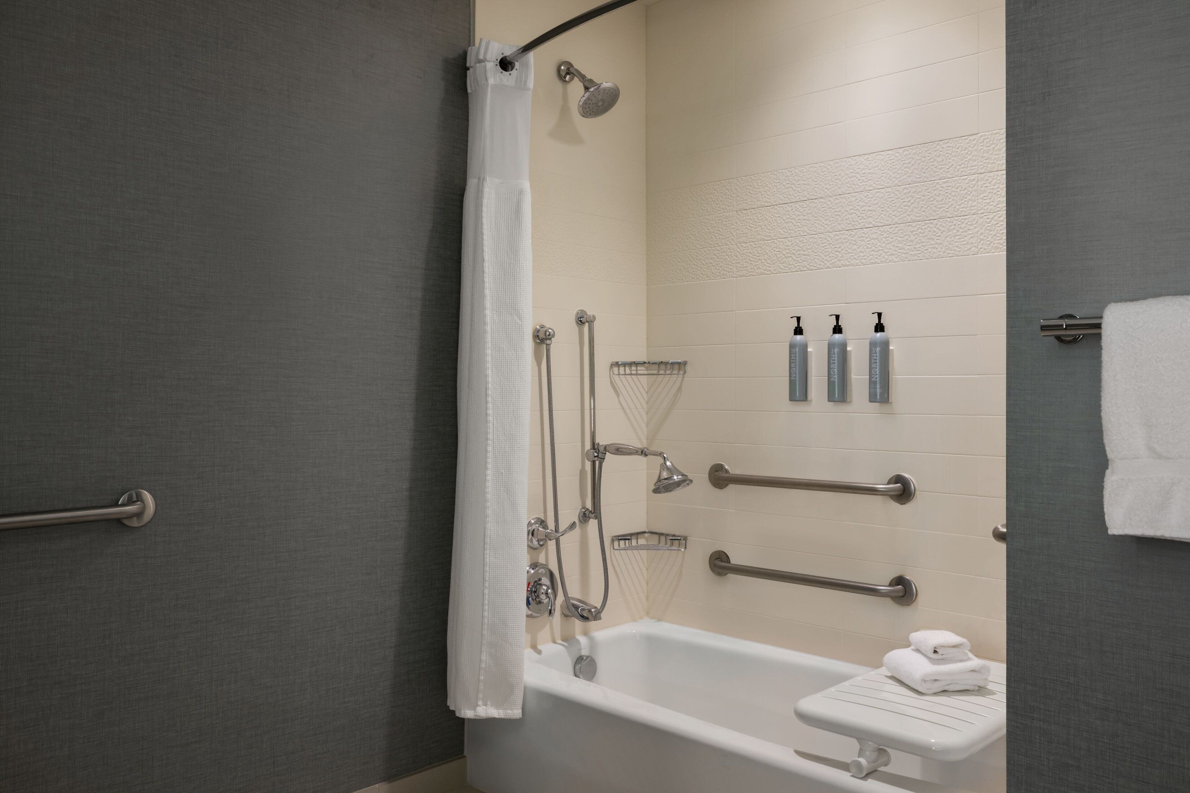 A modern bathroom with a bathtub, shower, and handrails. There are toiletries on the wall and a towel rack.