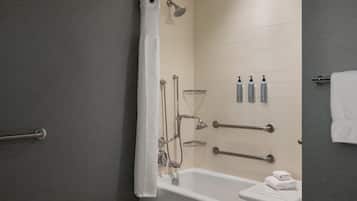 A modern bathroom with a bathtub, shower, and handrails. There are toiletries on the wall and a towel rack.