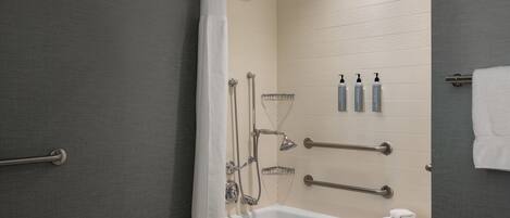 Un baño moderno con bañera, ducha y barras de apoyo. Hay artículos de aseo en la pared y un perchero para toallas.