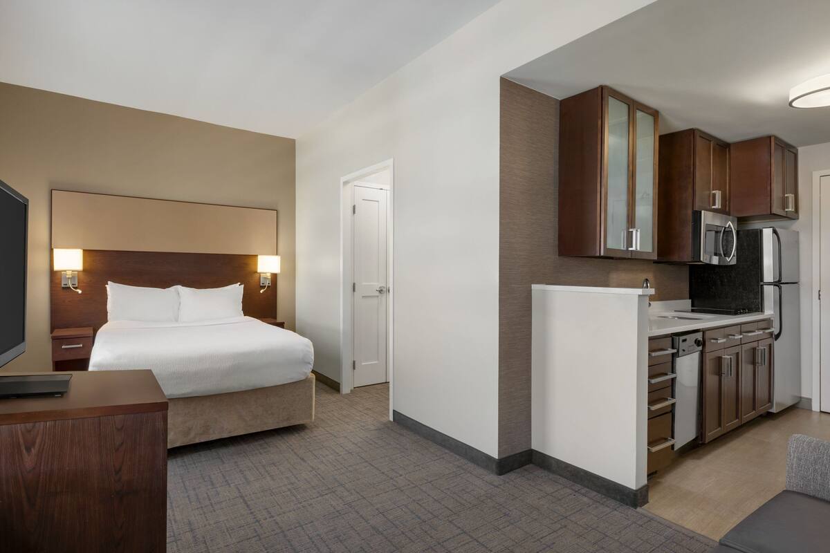 Een hotelkamer met een bed, nachtkastje, lamp en een kitchenette met een magnetron, koelkast en kastjes.