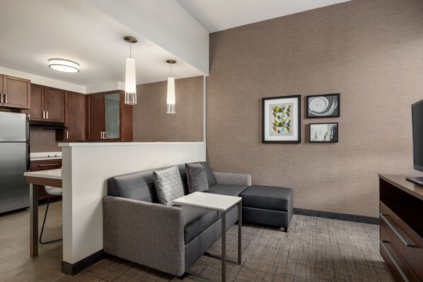 A modern kitchen with a refrigerator, a dining table, and a sofa. There are framed pictures on the wall and a pendant light above the sofa.