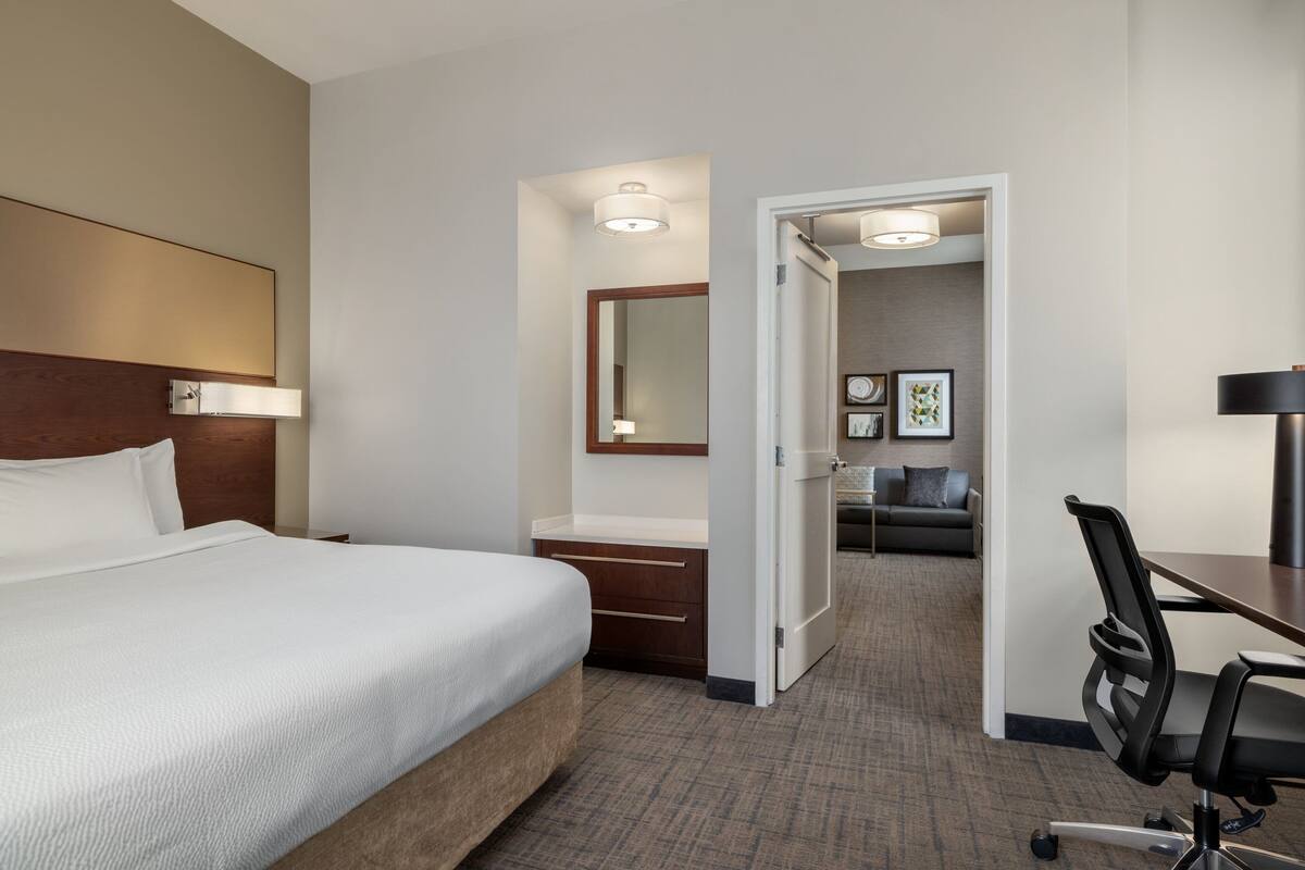 Een hotelkamer met een groot bed, een bureau met stoel, een dressoir en een spiegel. Er is een deur naar een zithoek met een bank en een salontafel.