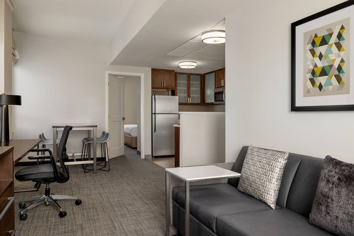 Een moderne hotelkamer met een grijze bank, een klein wit tafeltje, een zwarte bureaustoel, een kitchenette en een geometrisch kunstwerk in een lijst aan de muur.