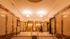 Un hall d’hôtel aux murs de marbre, doté d’un luminaire central et de deux ascenseurs.
