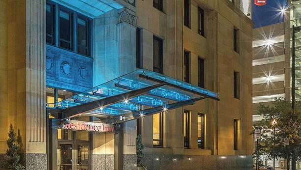 L’entrée d’un hôtel, avec un auvent illuminé, une grande porte vitrée et un tapis rouge.