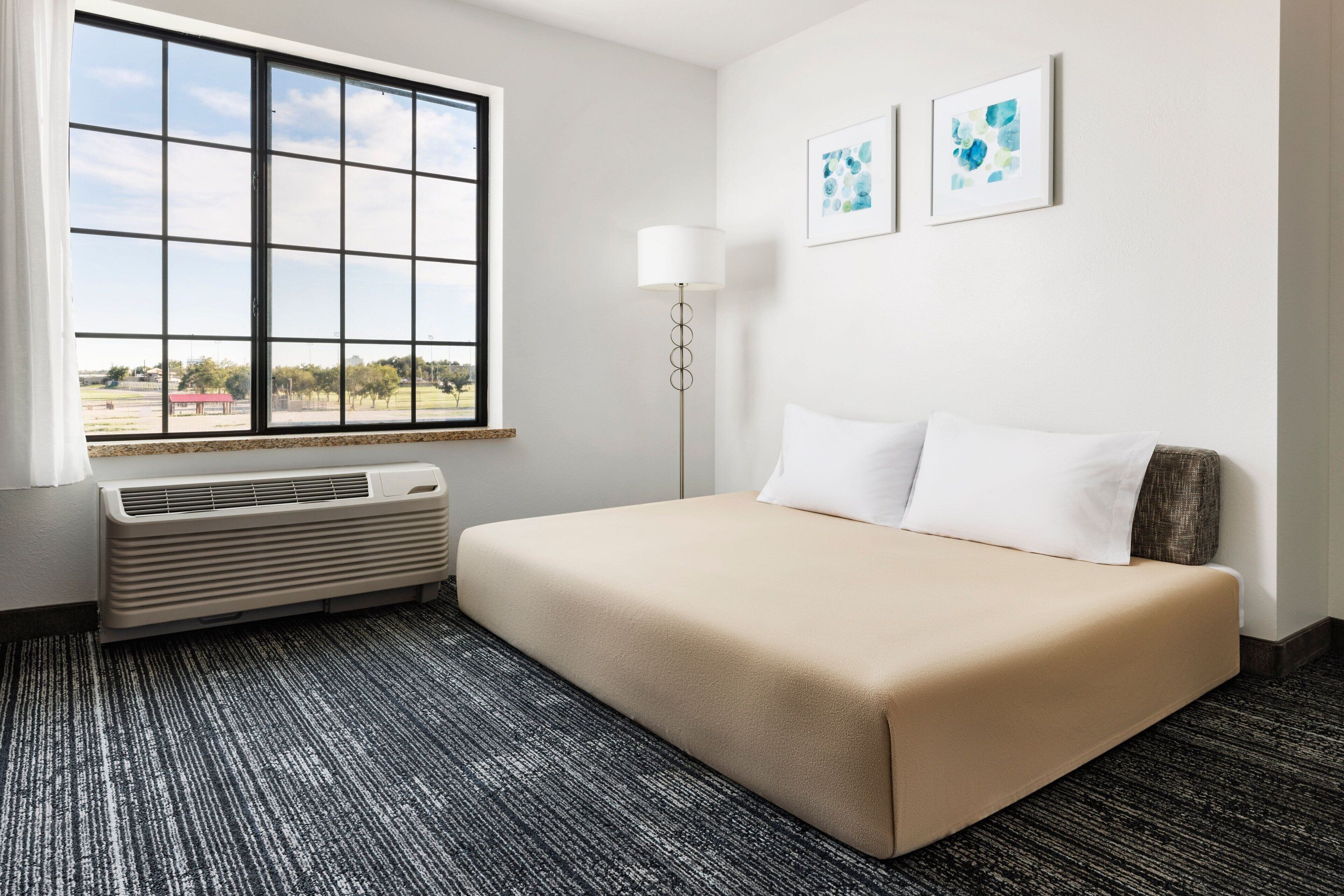 Habitación de hotel con un ventanal grande, una cama con ropa blanca, una lámpara de pie y dos cuadros en la pared.