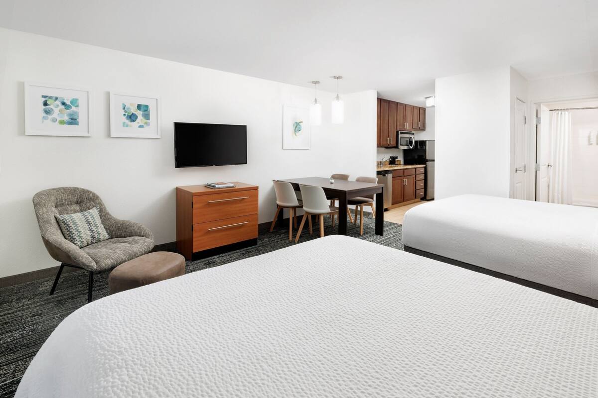 A hotel room with two beds, a desk, a chair, a television, and a kitchenette.